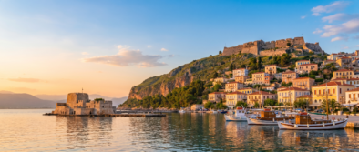 Sunset over a Mediterranean harbor: a stone fortress on a hillside overlooking pastel houses and moored boats by calm water on the shore point.